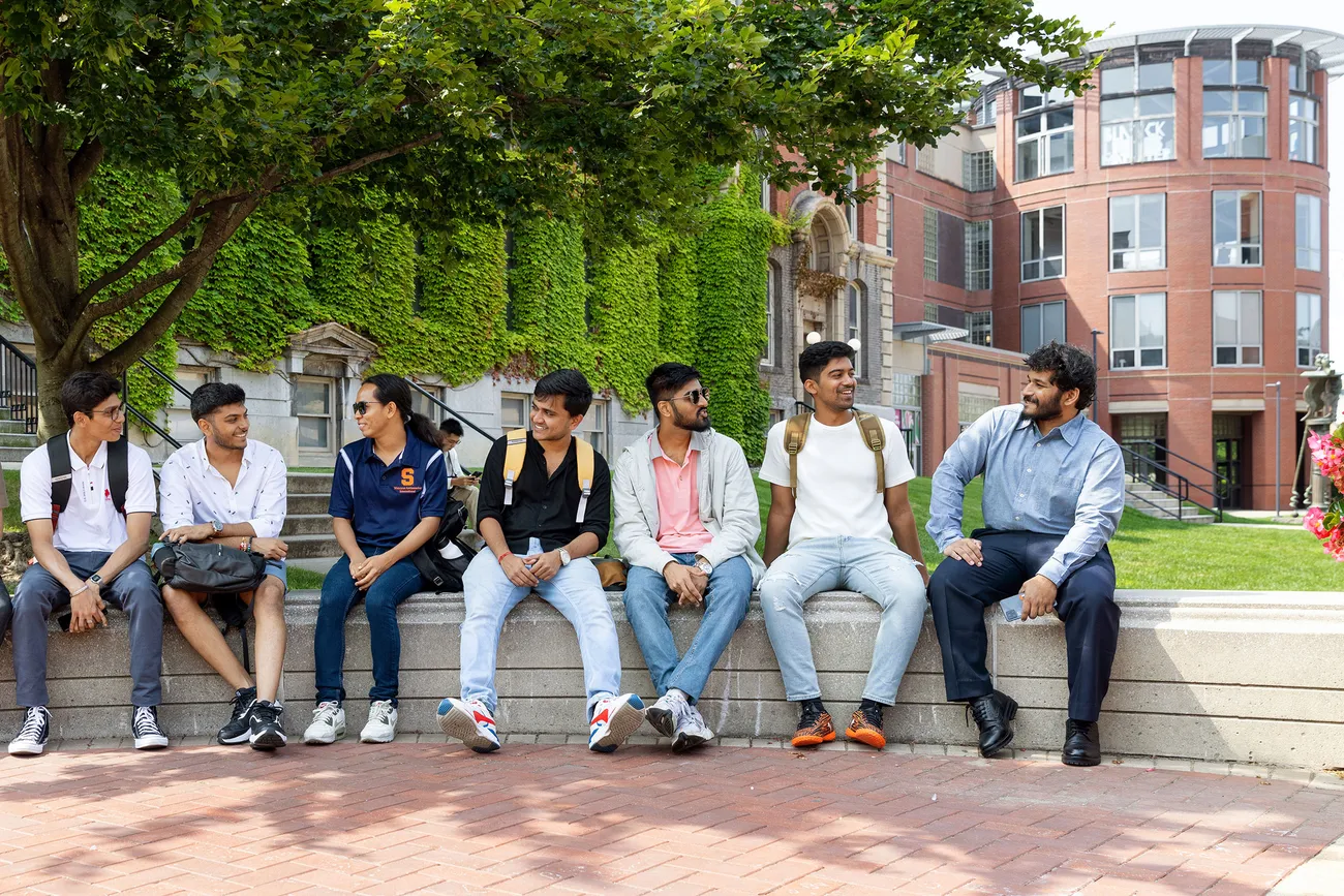 Seven students sitting on a bench waiting for a bus.