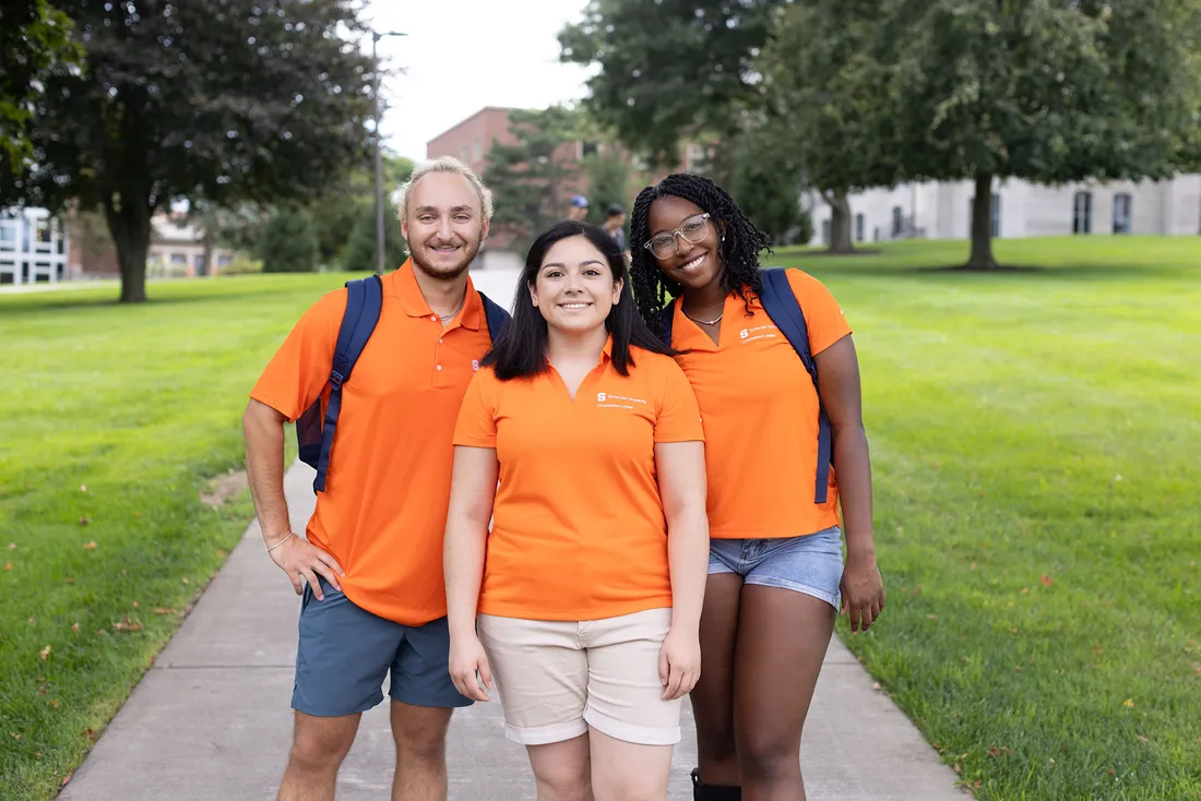 Three students in orange polo shirts standing together on Shaw Quadrangle.