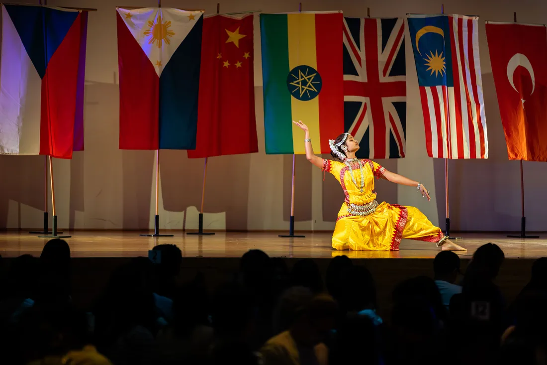 Student in traditional dress performs on stage with international flags in background.